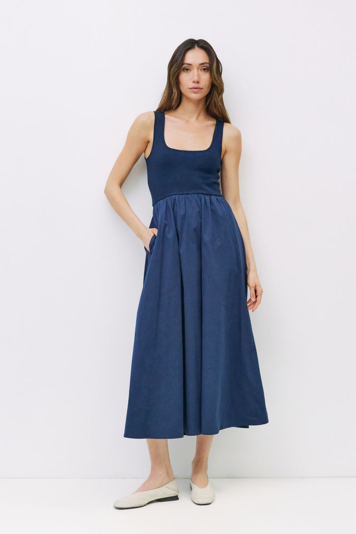 woman wearing a navy blue sleeveless maxi dress with a fitted bodice and gathered skirt, standing with hands in pockets