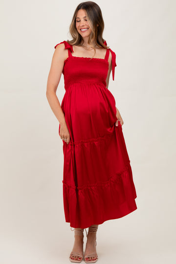 woman wearing a red maternity maxi dress with tie straps and a tiered skirt, gently holding her baby bump