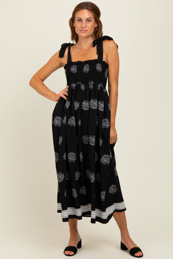 woman wearing a black midi dress with white floral patterns and tie shoulder straps, standing with one hand on her hip