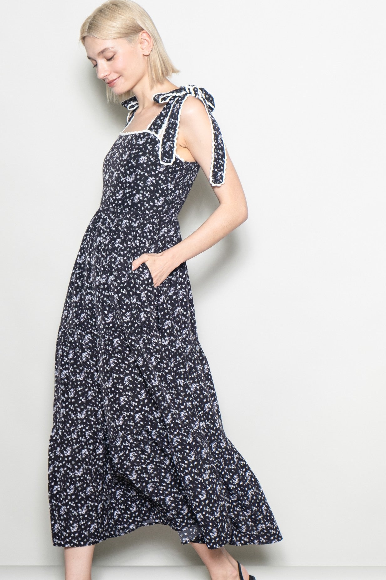 woman wearing a navy blue floral maxi dress with bow tie straps and side pockets, standing sideways