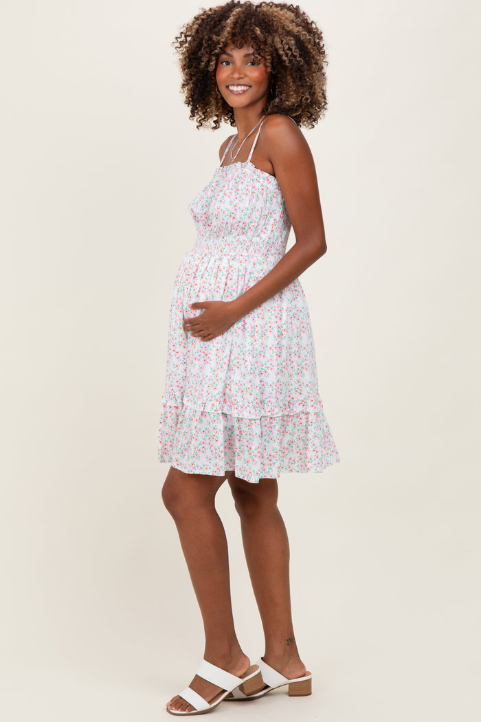 woman wearing a white floral maternity dress with spaghetti straps and a baby bump visible, smiling and holding her belly