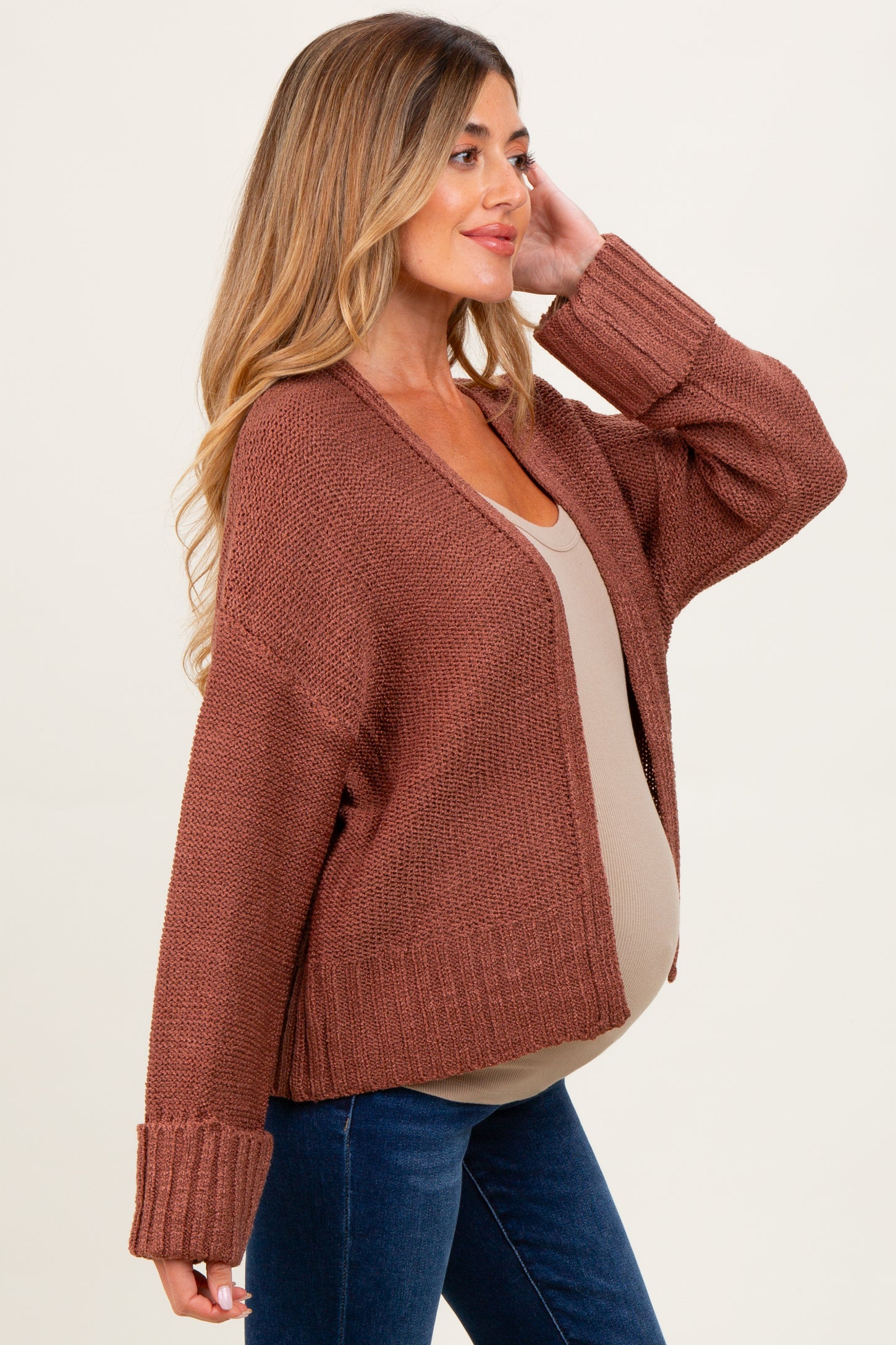 woman wearing a rust-colored open-knit maternity cardigan over a taupe top and showing her baby bump, styled with jeans