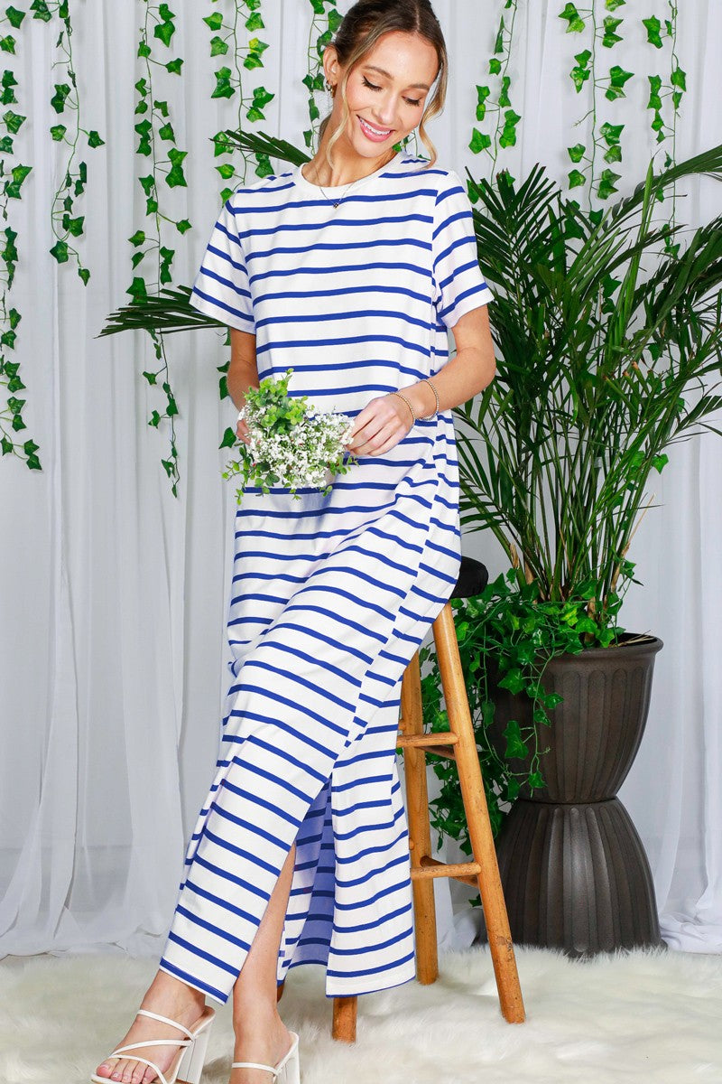 woman wearing a white and navy striped maxi dress with side slits, standing next to a plant and holding a bouquet of flowers