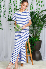 woman wearing a white and navy striped maxi dress with side slits, standing next to a plant and holding a bouquet of flowers