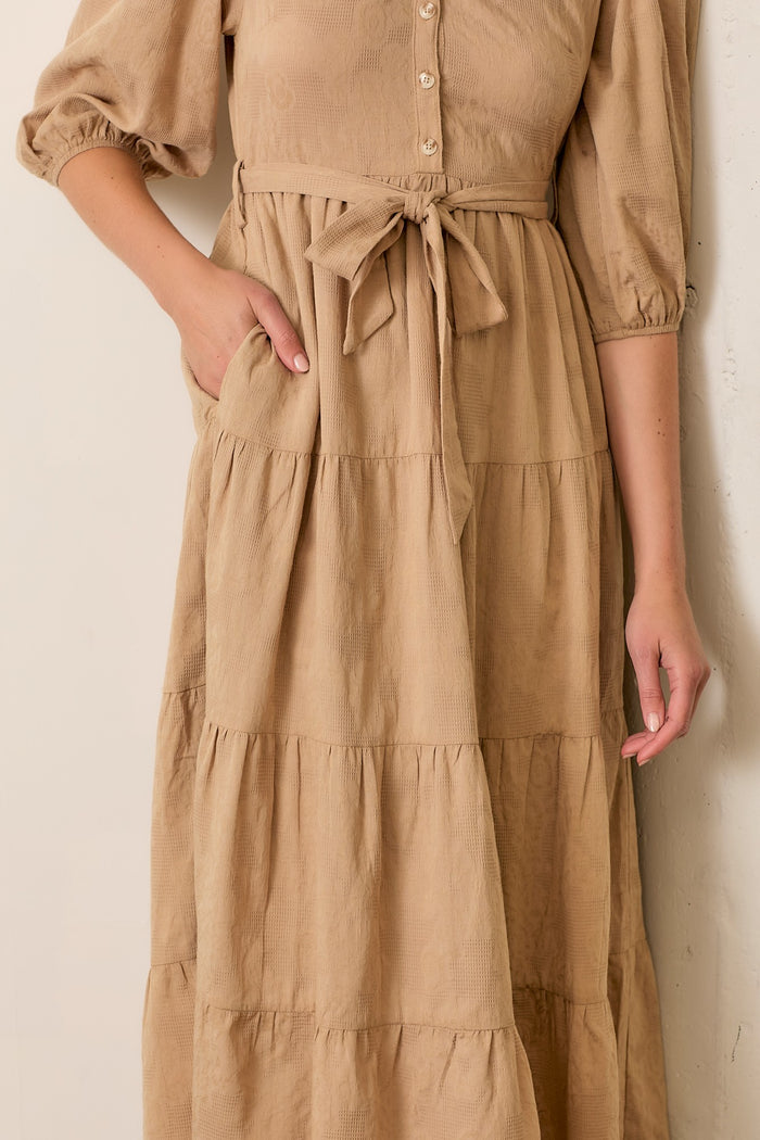woman wearing a beige tiered maxi dress with short puff sleeves and a tie waist detail, hand in pocket