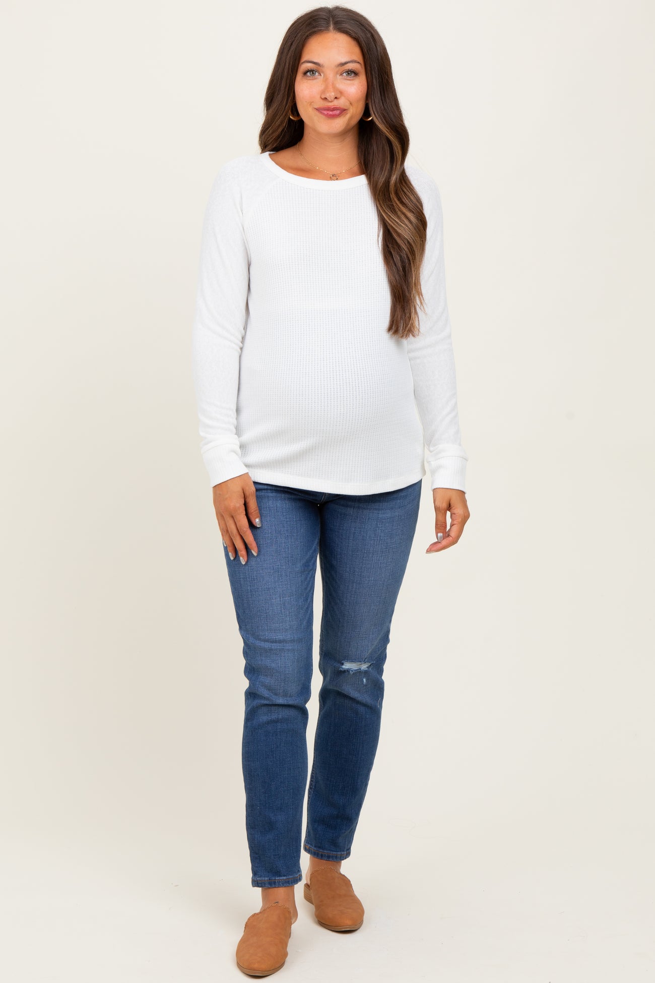 woman wearing a white long sleeve ribbed maternity top and blue distressed maternity jeans, showcasing her baby bump