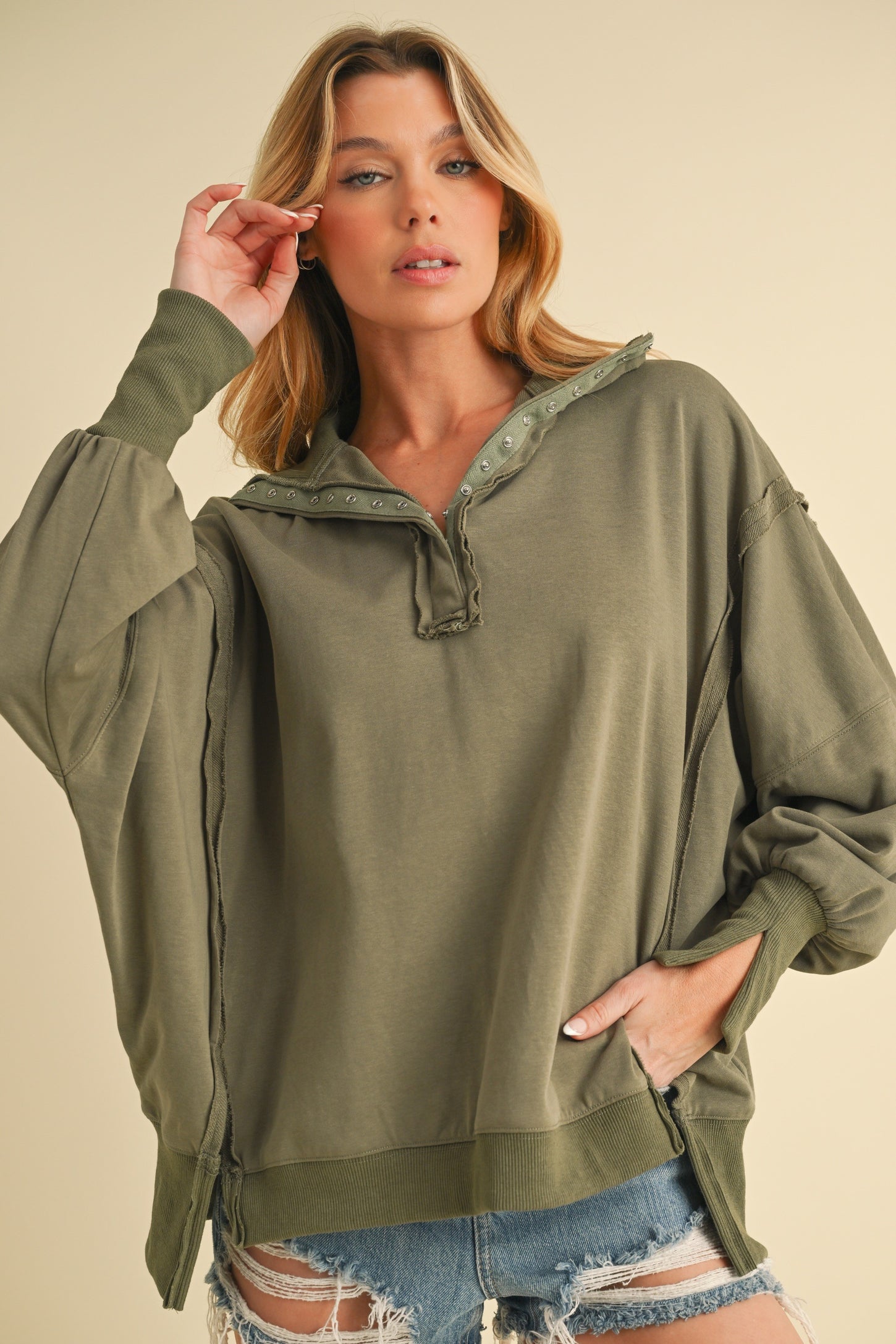 woman wearing an oversized olive green pullover with button details and side slits, styled with distressed denim shorts