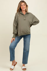 woman wearing an olive green maternity pullover sweatshirt and light blue jeans, smiling and standing against a neutral background
