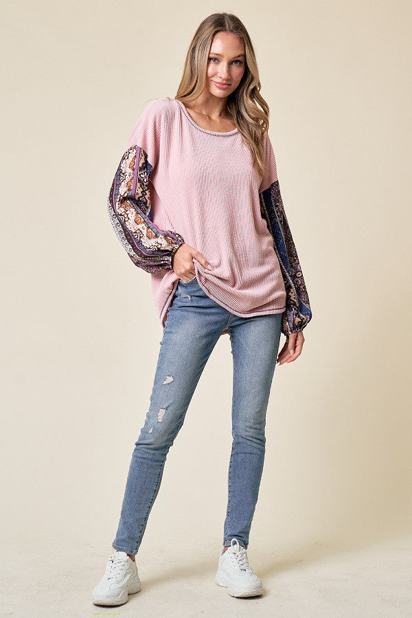woman wearing a pink ribbed long sleeve top with patterned sleeves and blue distressed jeans, standing on a beige background