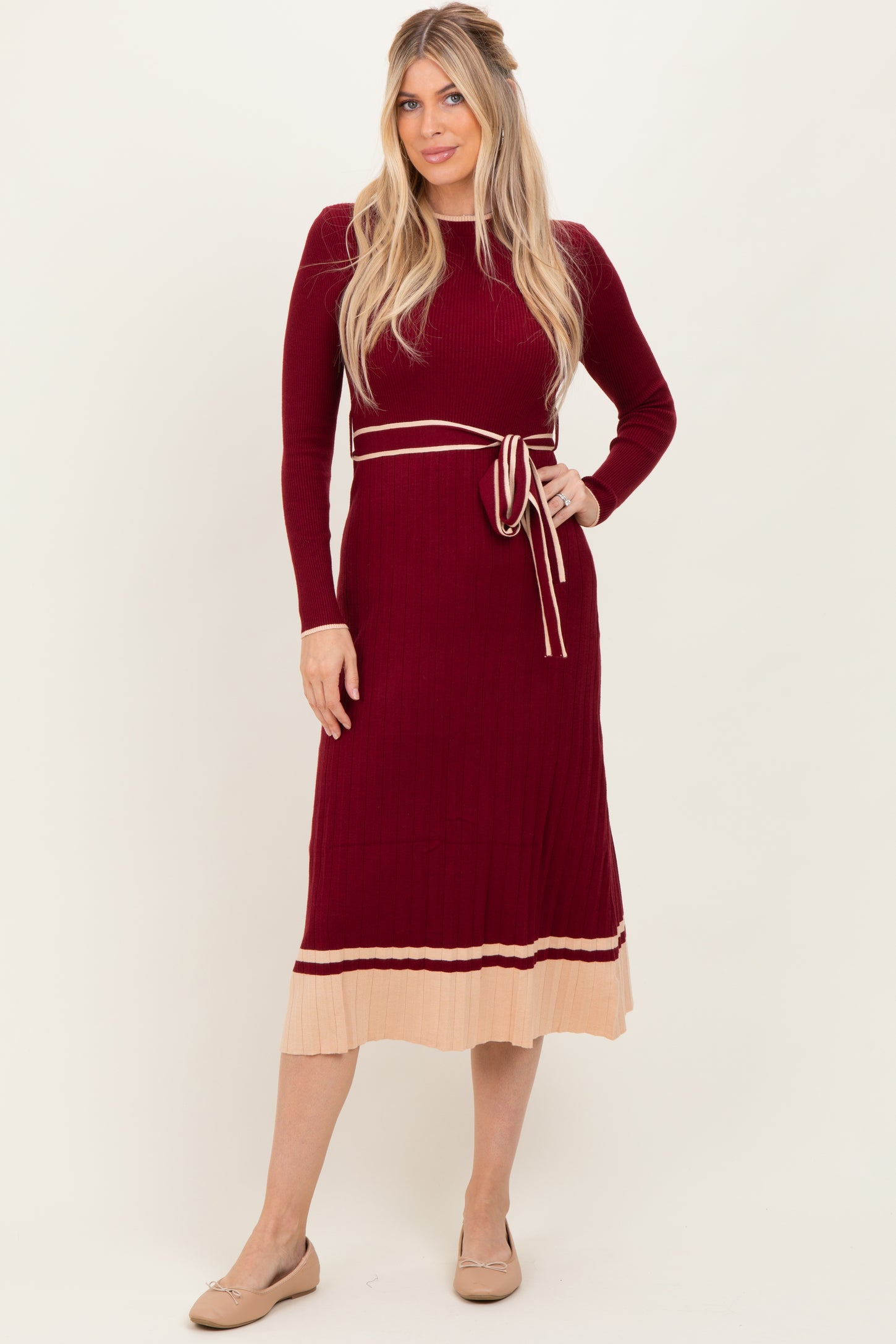 woman wearing a burgundy ribbed long sleeve dress with a tie belt and a beige pleated hem