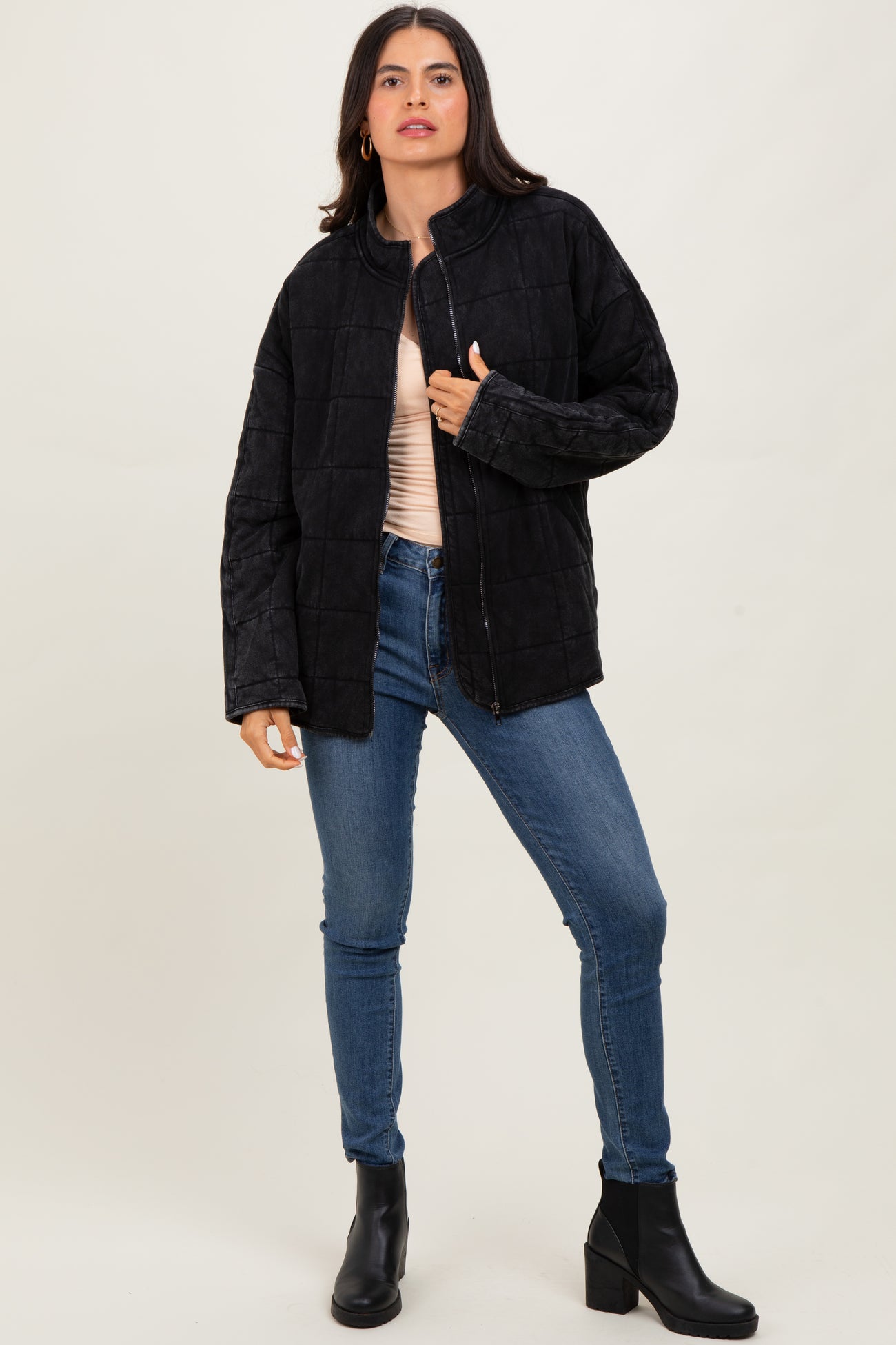 woman wearing a black quilted jacket, light pink top, and blue jeans standing in front of a neutral background