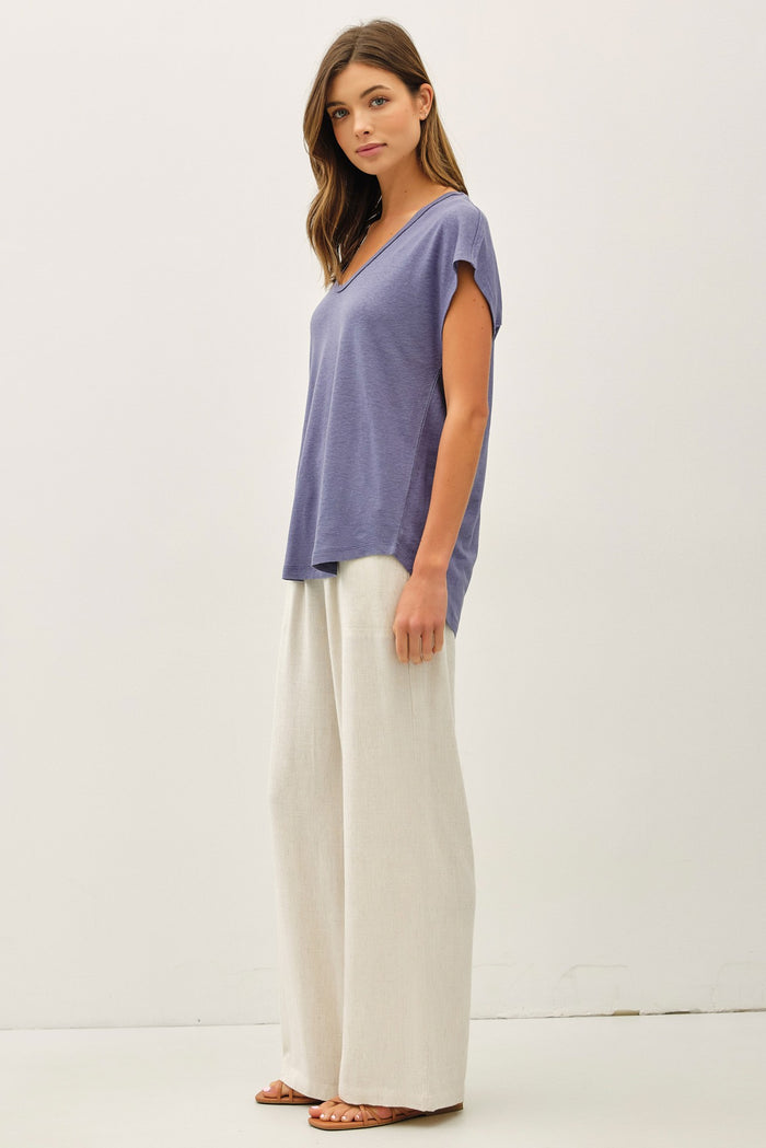 woman wearing a loose-fitting purple v-neck top and wide-leg cream pants, standing sideways against a neutral background