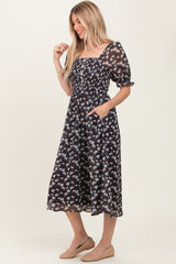 woman wearing a black floral midi dress with puff sleeves and a square neckline, featuring a fitted bodice and side pockets