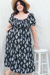 woman wearing a black floral print plus-size dress with puff sleeves and a cinched waist, smiling while standing beside a stool