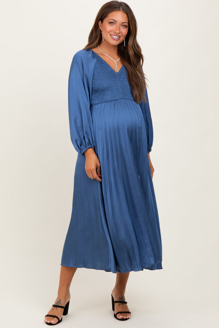 woman wearing a blue pleated maternity dress with a smocked bodice and long sleeves, showcasing her baby bump