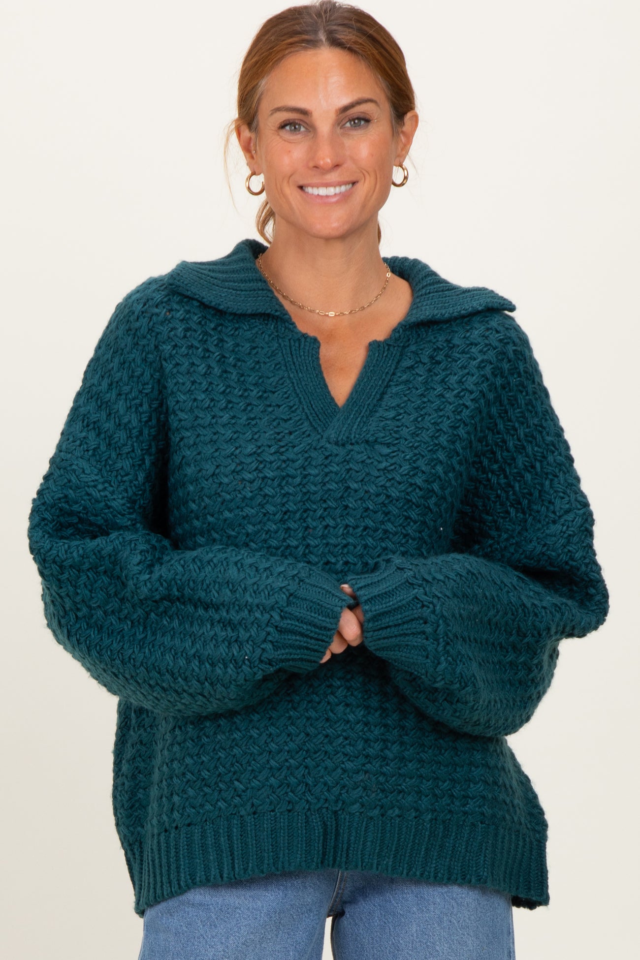 woman wearing a teal oversized knitted sweater with a v-neck and a collar, smiling and posing with hands clasped