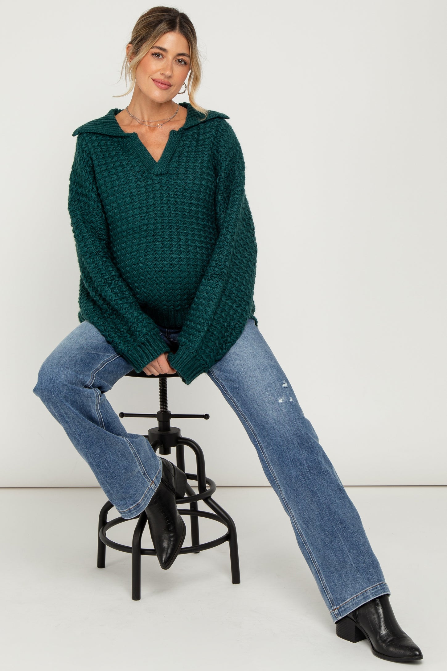 woman wearing a dark green chunky knit maternity sweater and blue distressed jeans, highlighting her baby bump