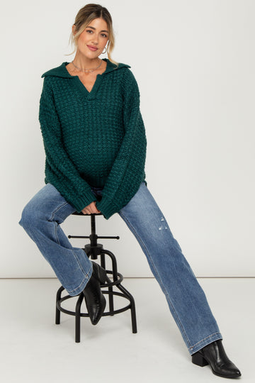woman wearing a dark green chunky knit maternity sweater and blue distressed jeans, highlighting her baby bump