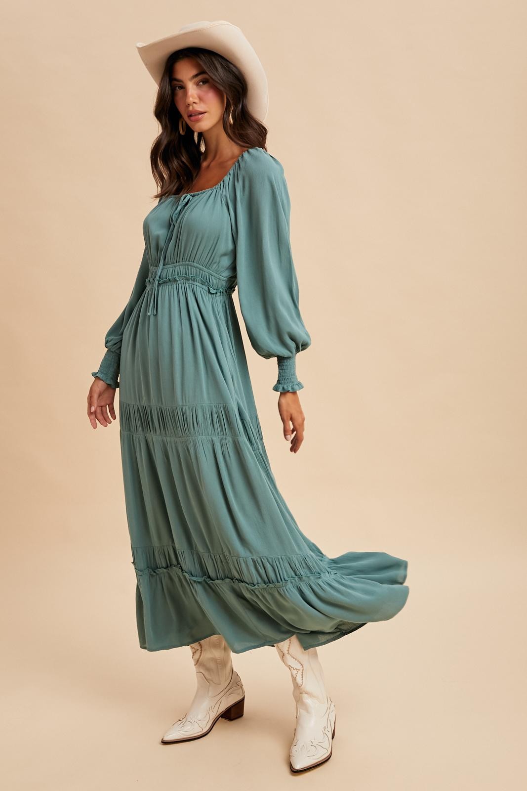 woman wearing a teal long sleeve maxi dress with ruffle details and white cowboy boots, standing against a neutral background