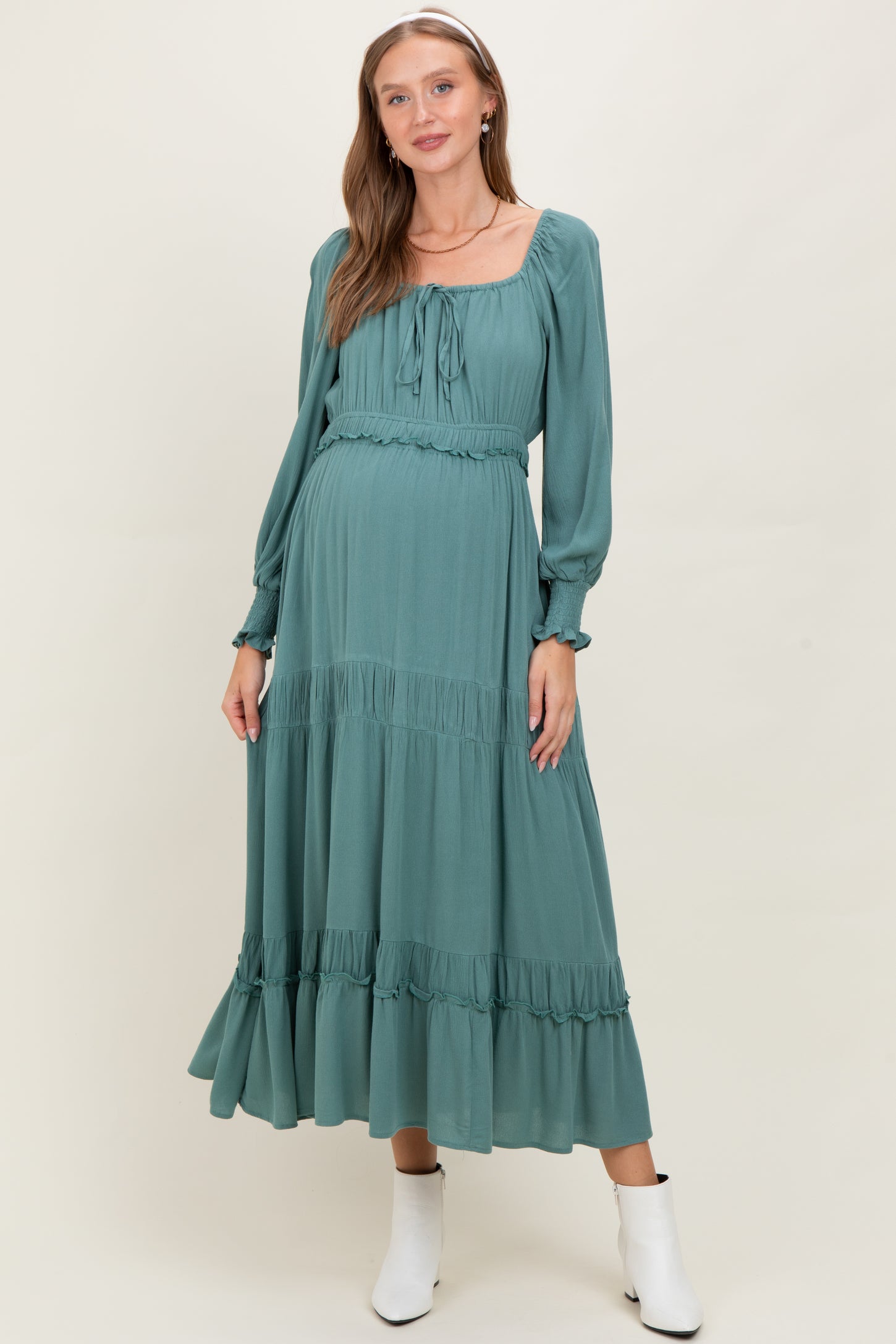 woman wearing a dusty teal maternity dress with long sleeves and tiered ruffle details, featuring a tied neckline