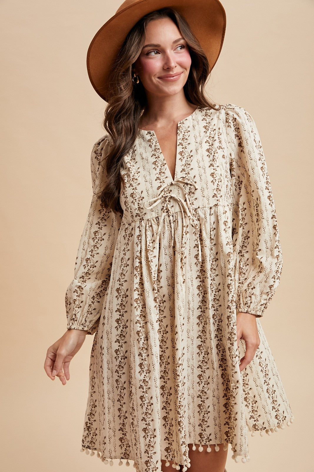 woman wearing a cream floral print long sleeve dress with a v-neck and pom pom hem, accessorized with a wide-brimmed hat