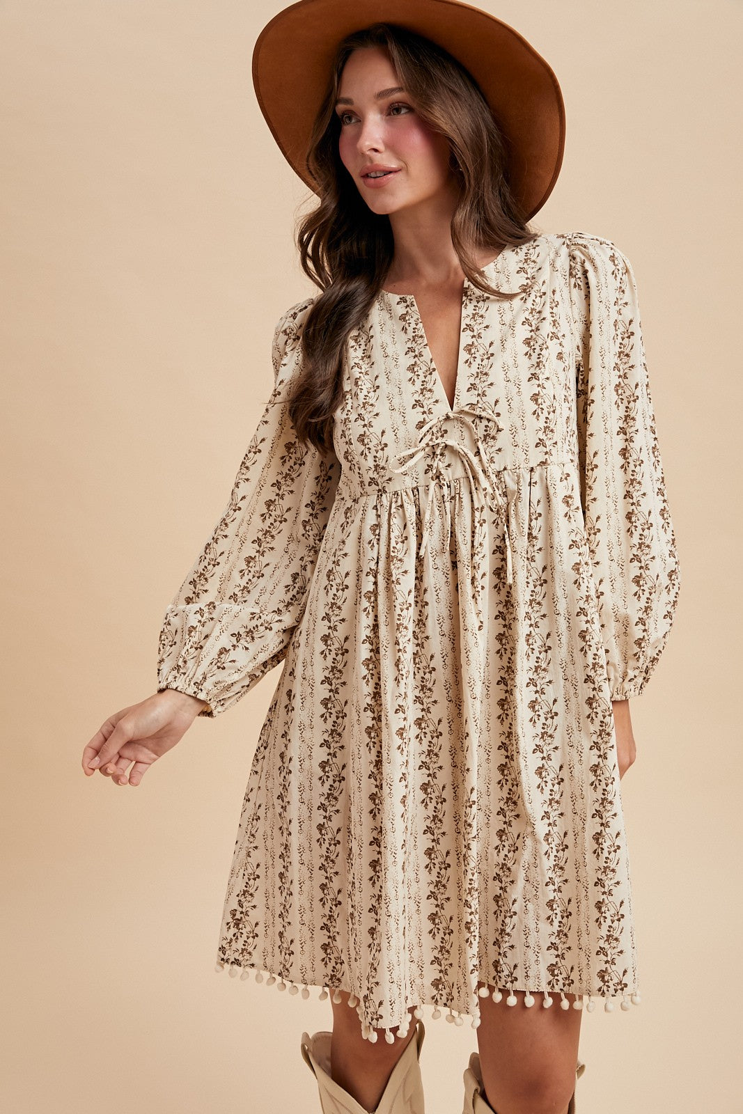 woman wearing a cream floral long sleeve dress with a tie detail and pom-pom trim, accessorized with a brown hat