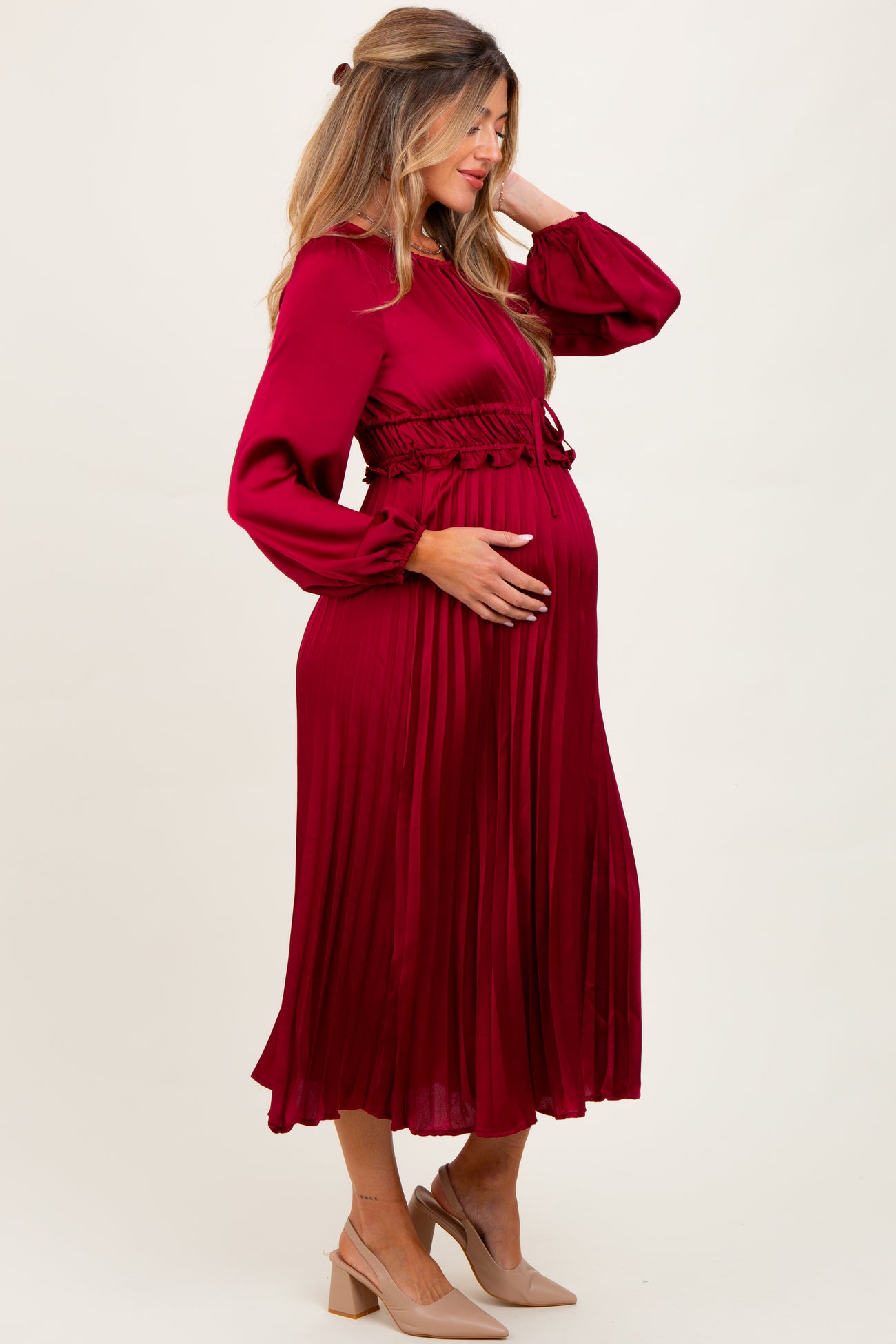 woman wearing a burgundy pleated maternity dress with long sleeves and a fitted waist, holding her baby bump