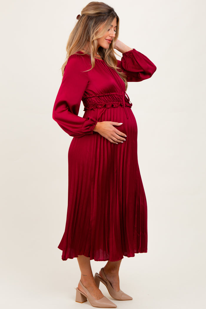 woman wearing a burgundy pleated maternity dress with long sleeves and a fitted waist, holding her baby bump