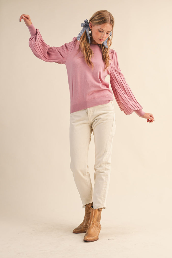 woman wearing a light pink sweater with pleated sleeves and white jeans, standing in a relaxed pose