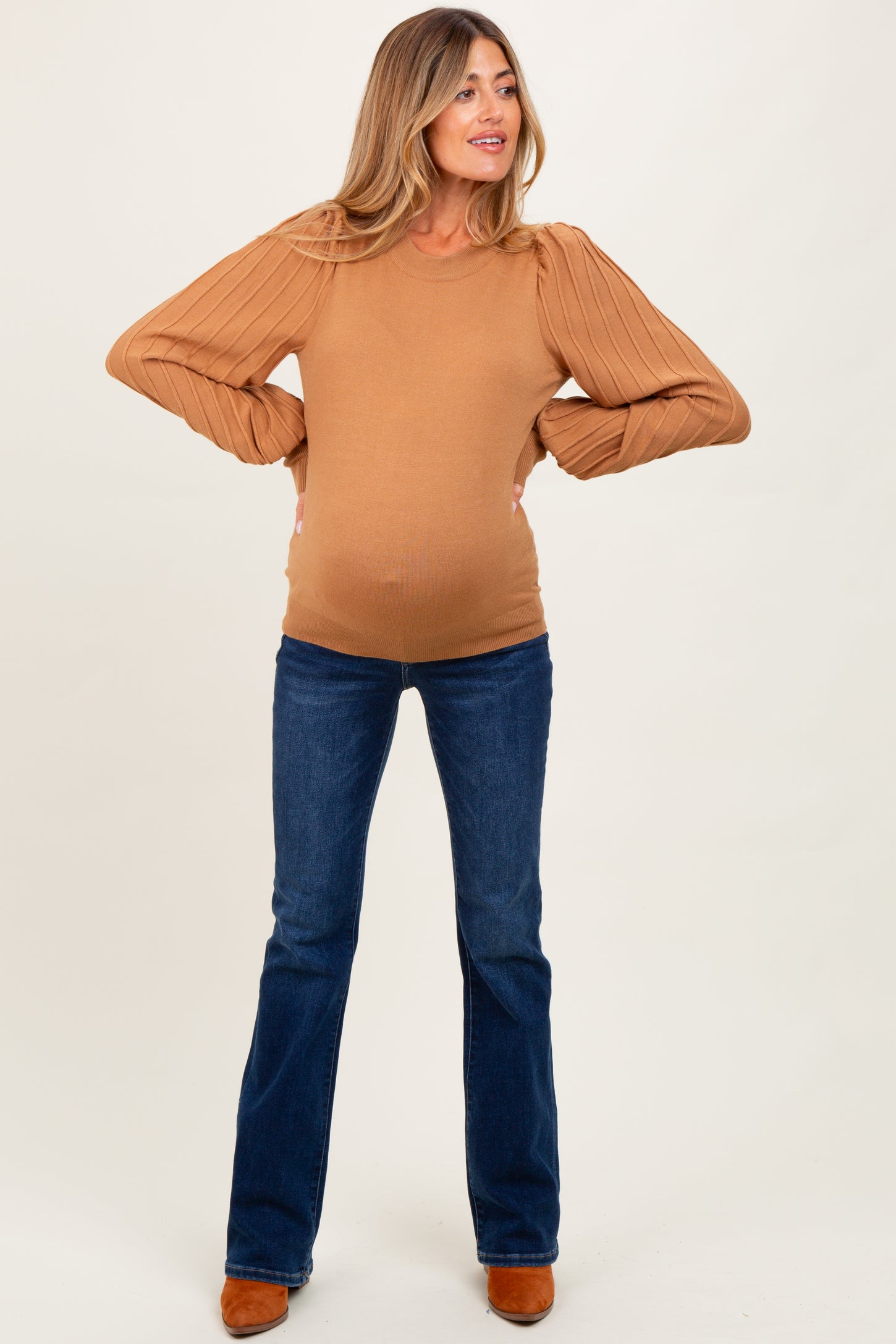 woman wearing a camel-colored maternity sweater with puffed sleeves and blue flared jeans, showing her baby bump