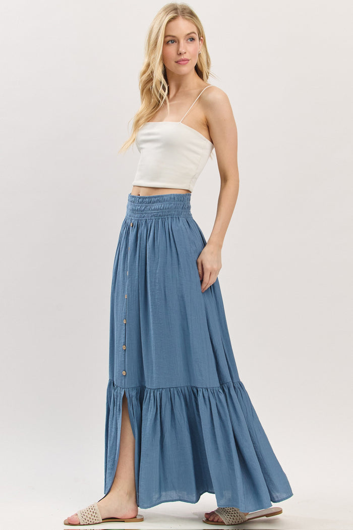 woman wearing a light blue maxi skirt with a tiered hem and button details, paired with a white crop top and sandals