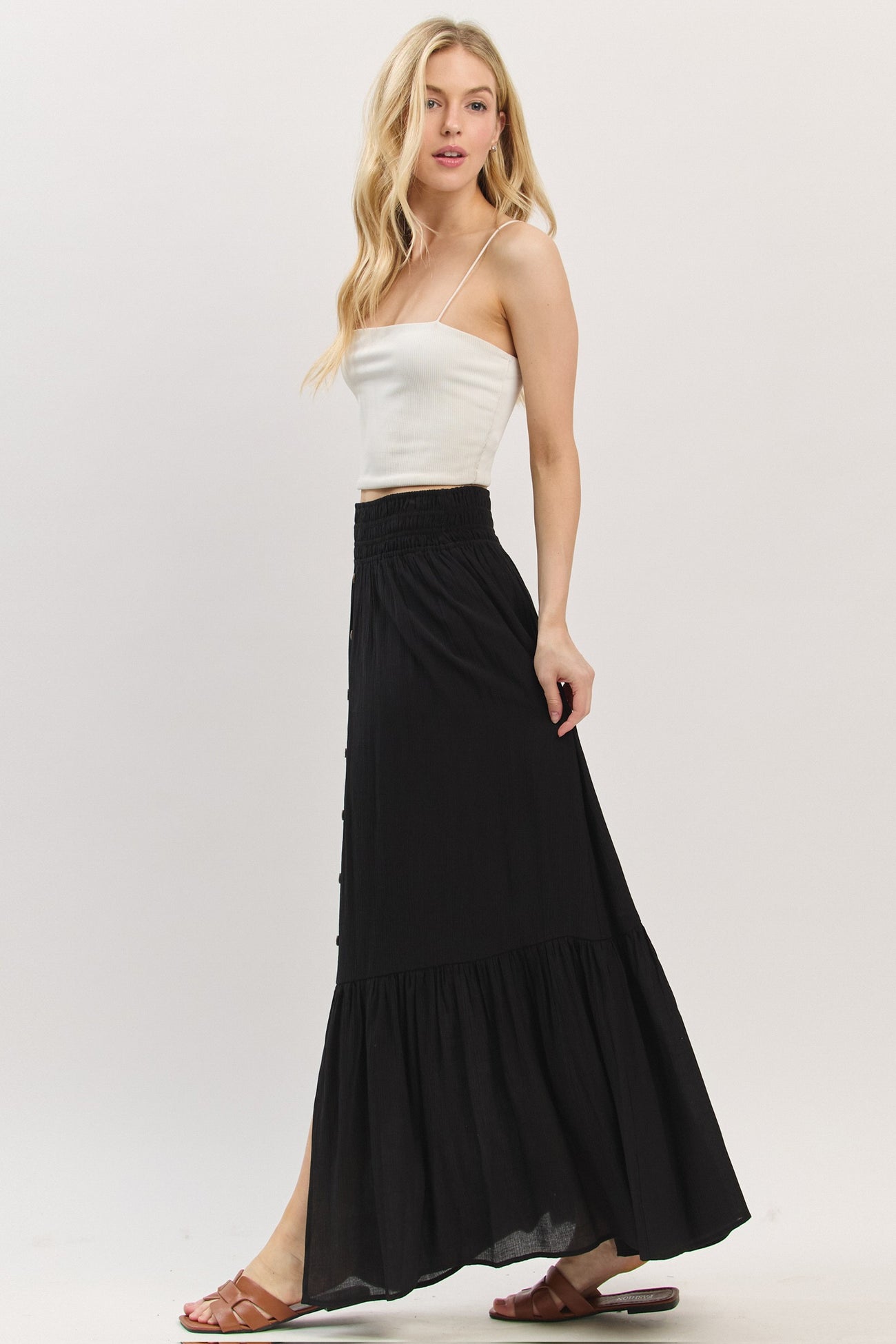 woman wearing a white ribbed tank top and a black tiered maxi skirt with a button detail and side slit