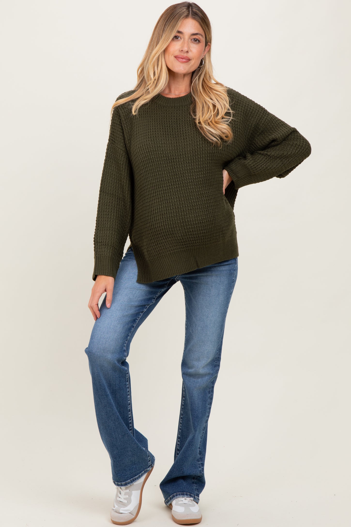 woman wearing a dark green textured maternity sweater and blue flared jeans, showing her baby bump