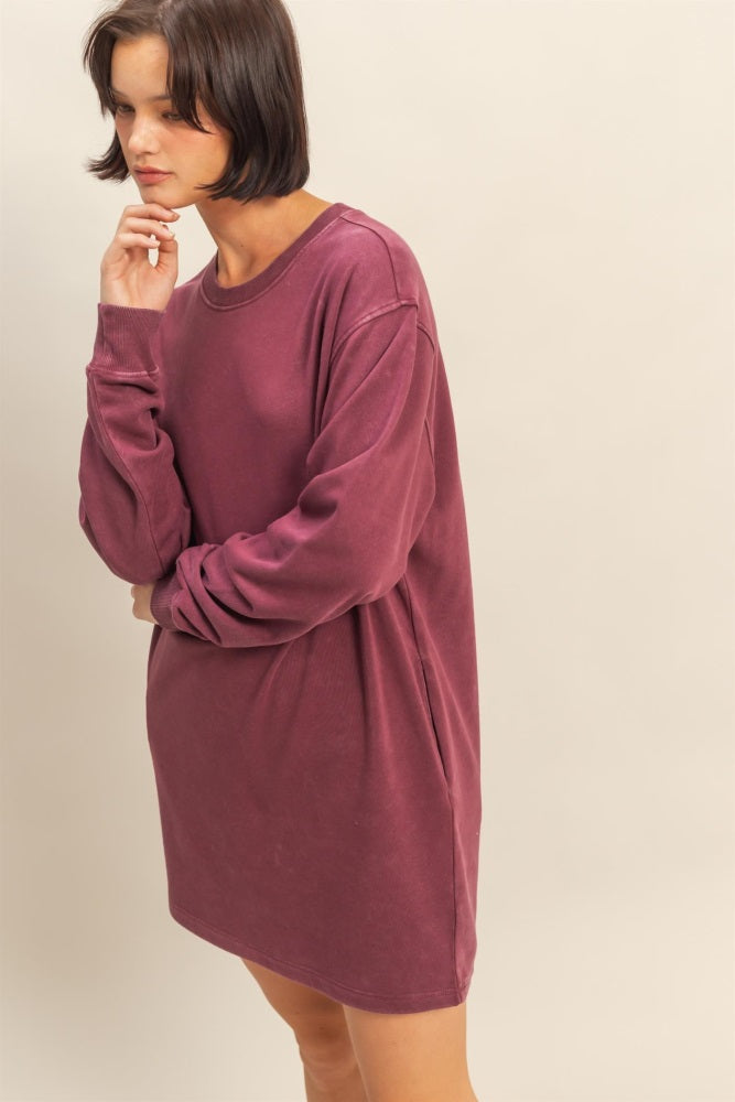 woman wearing a long-sleeve oversized burgundy sweatshirt dress standing with one hand on her chin