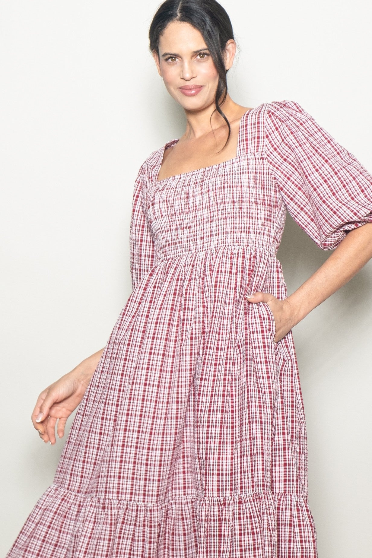 woman wearing a red and white checkered puff sleeve dress with a square neckline and tiered hem, posing with one hand in pocket