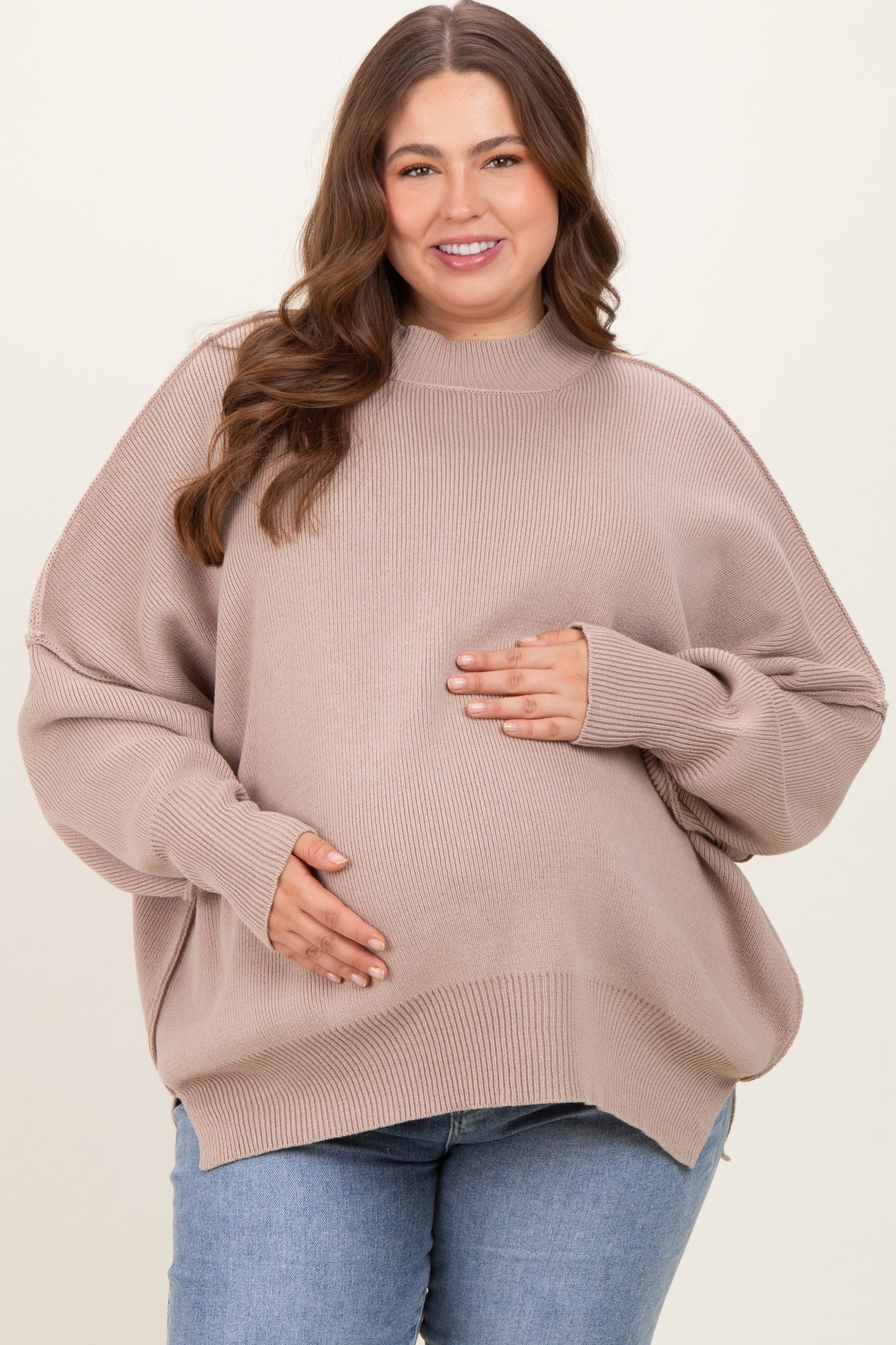 woman wearing a beige oversized maternity sweater and holding her baby bump, paired with blue jeans