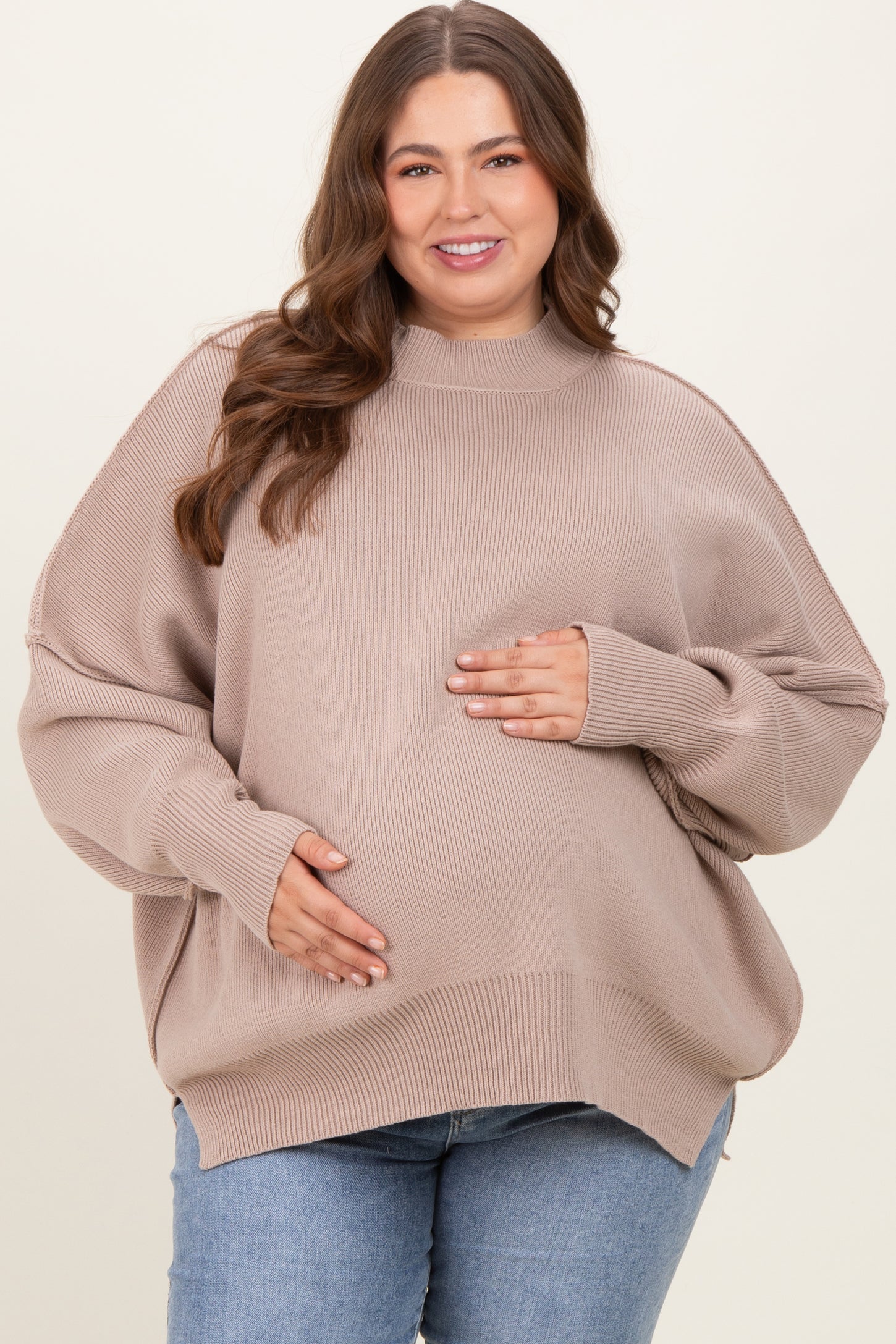 woman wearing a beige oversized maternity sweater and holding her baby bump, paired with blue jeans