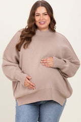 woman wearing a beige oversized maternity sweater and holding her baby bump, paired with blue jeans