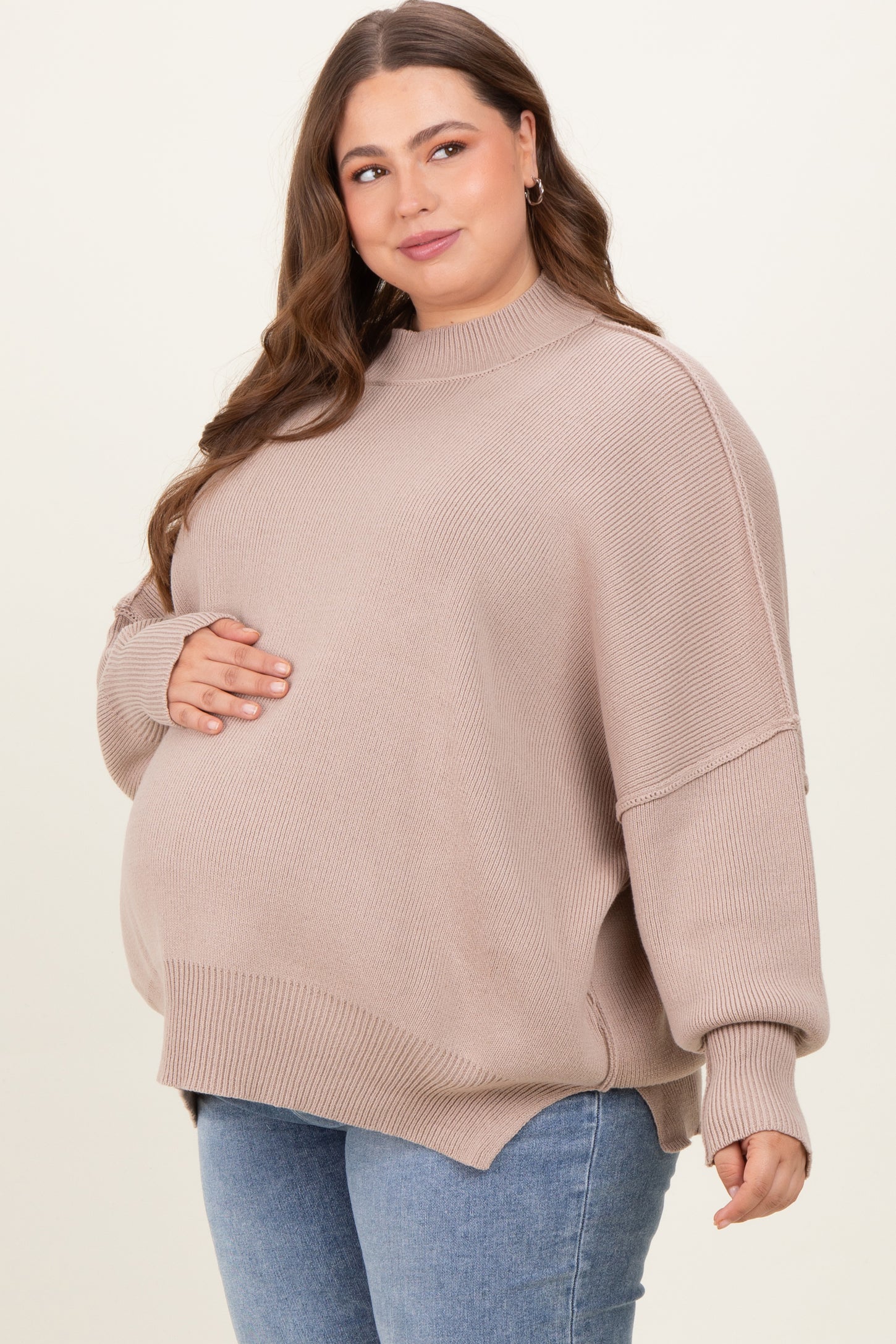 woman wearing a beige oversized maternity sweater, cradling her baby bump, paired with blue jeans