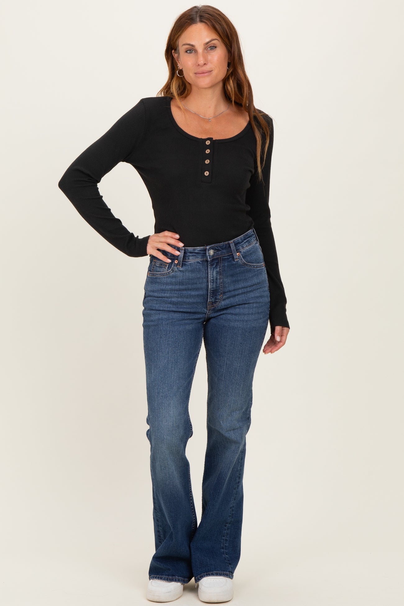 woman wearing a black long sleeve ribbed top with buttons and blue flared jeans standing upright