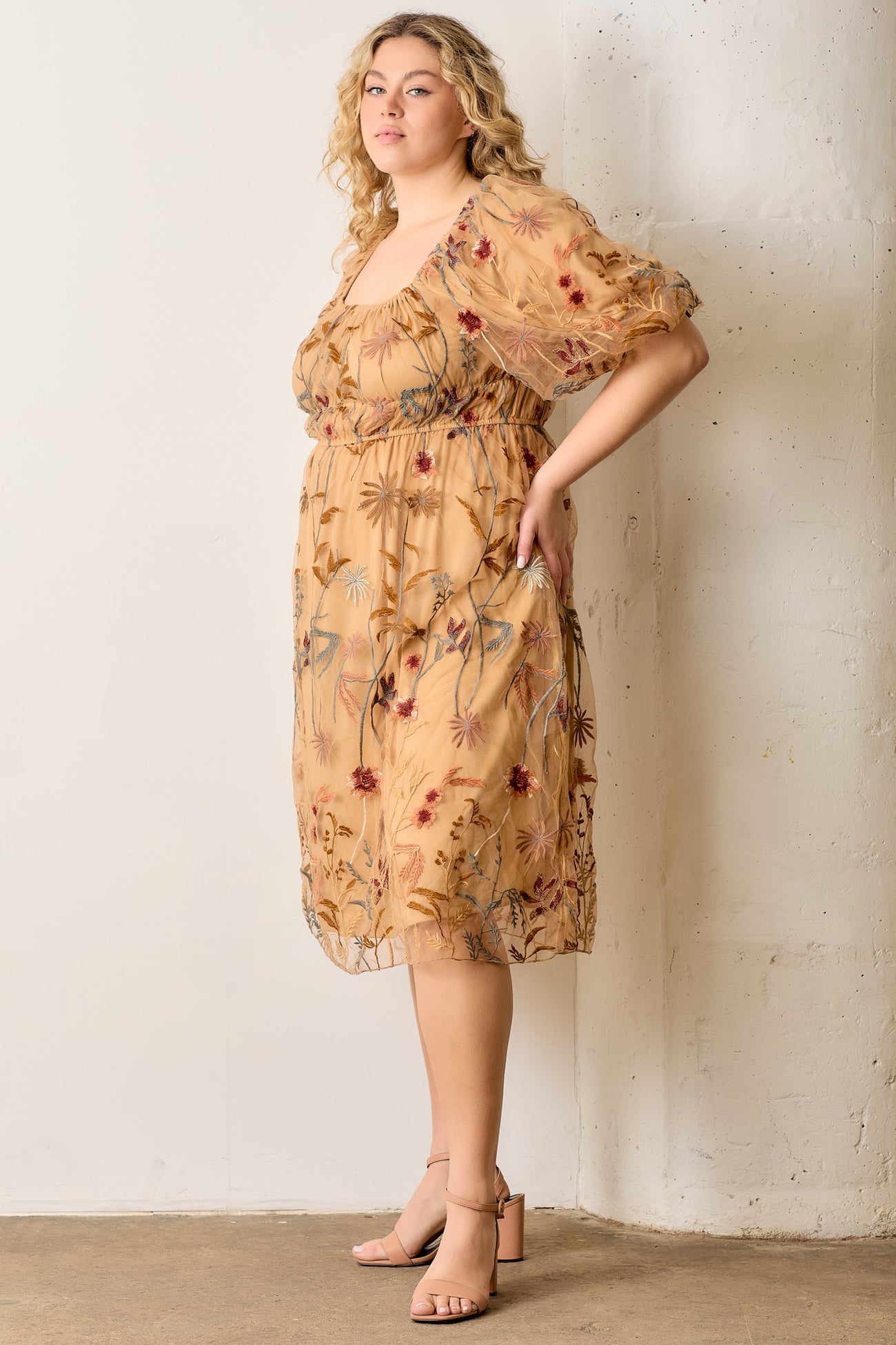 woman wearing a floral embroidered plus size dress with puff sleeves and a square neckline, standing against a wall