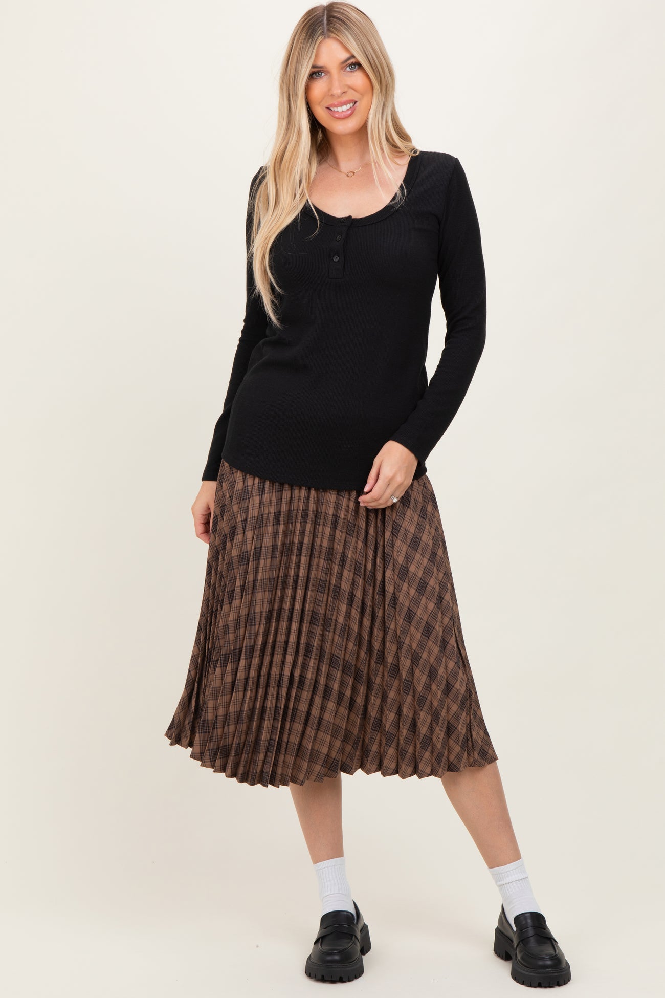 woman wearing a black long sleeve henley top with a brown pleated checked midi skirt and black chunky loafers