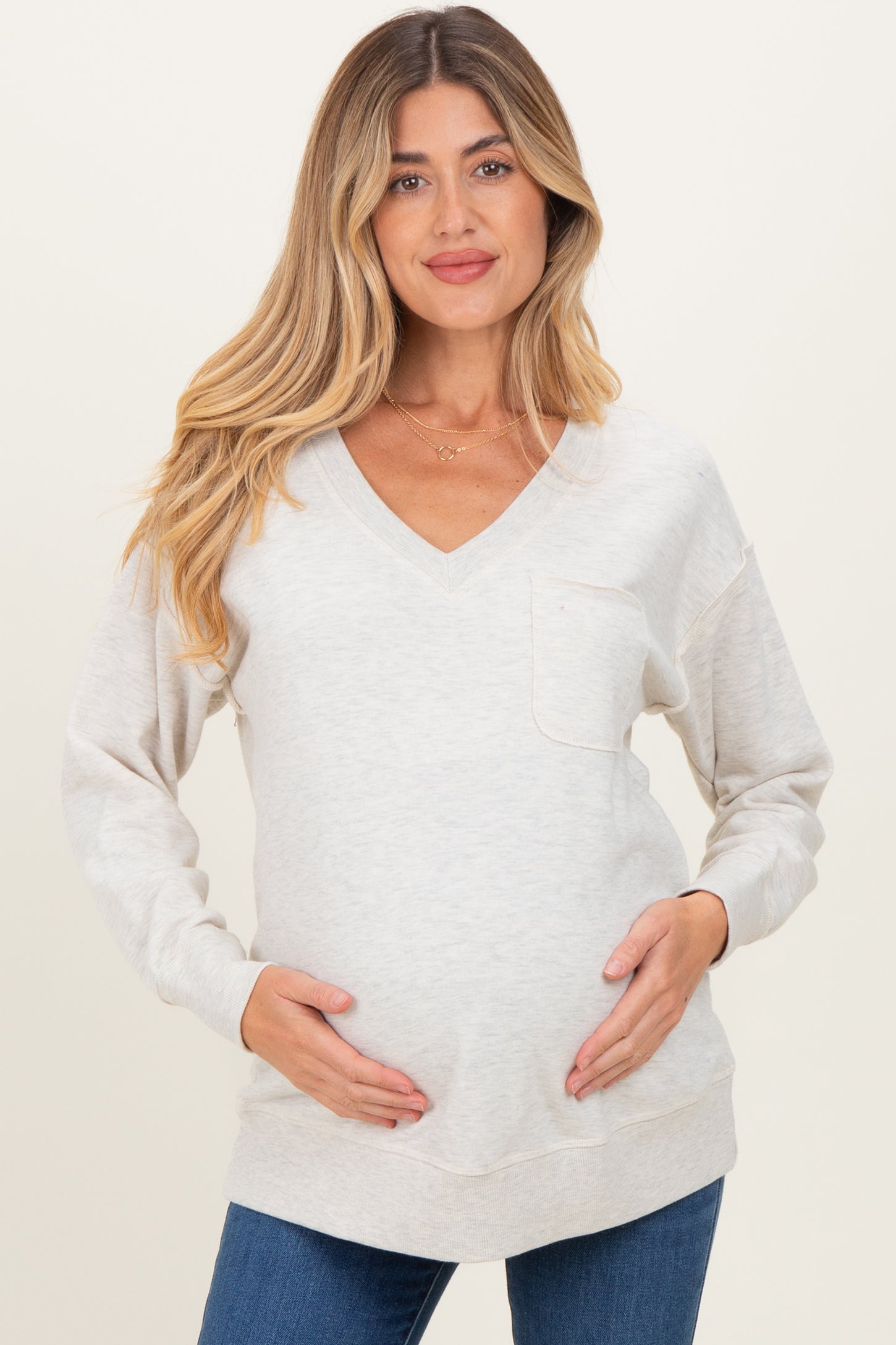 woman wearing a light gray maternity sweatshirt with a v-neck and pocket, holding her baby bump