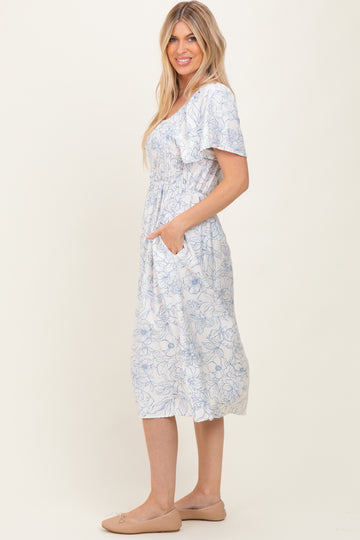 woman wearing a light blue floral midi dress with smocked bodice and short sleeves, standing with hands in pockets