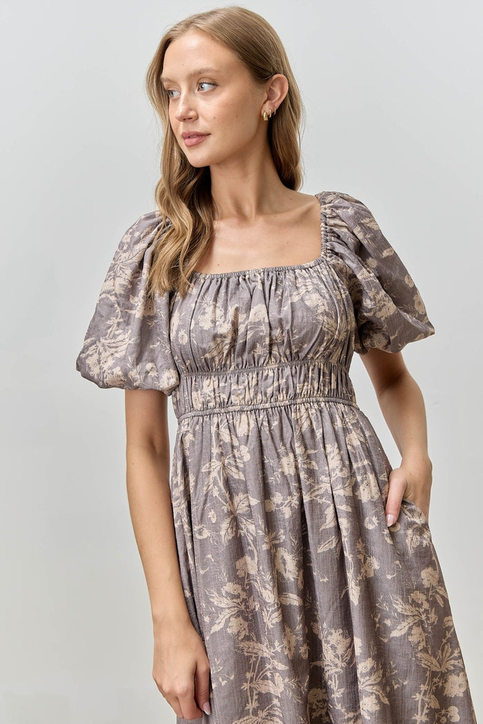woman wearing a brown floral puff sleeve dress with a square neckline and elastic waist, posing with hand in pocket