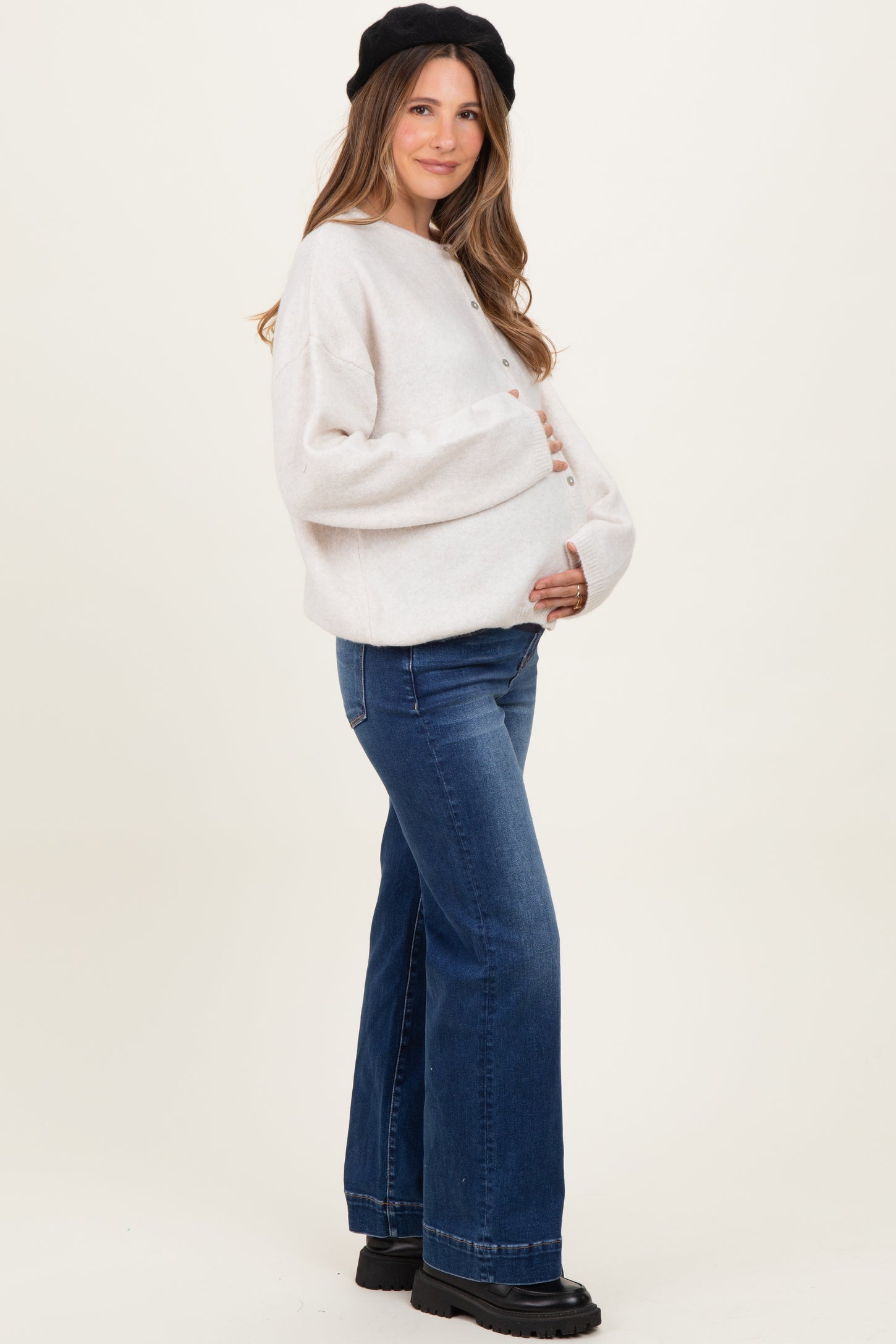 woman wearing a cream oversized maternity sweater and flared jeans, holding her baby bump