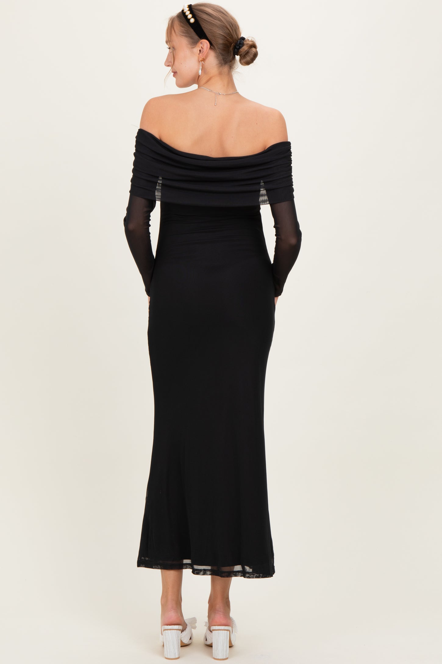 woman wearing a black off-shoulder maternity dress with ruched detailing and long sheer sleeves, showing no baby bump