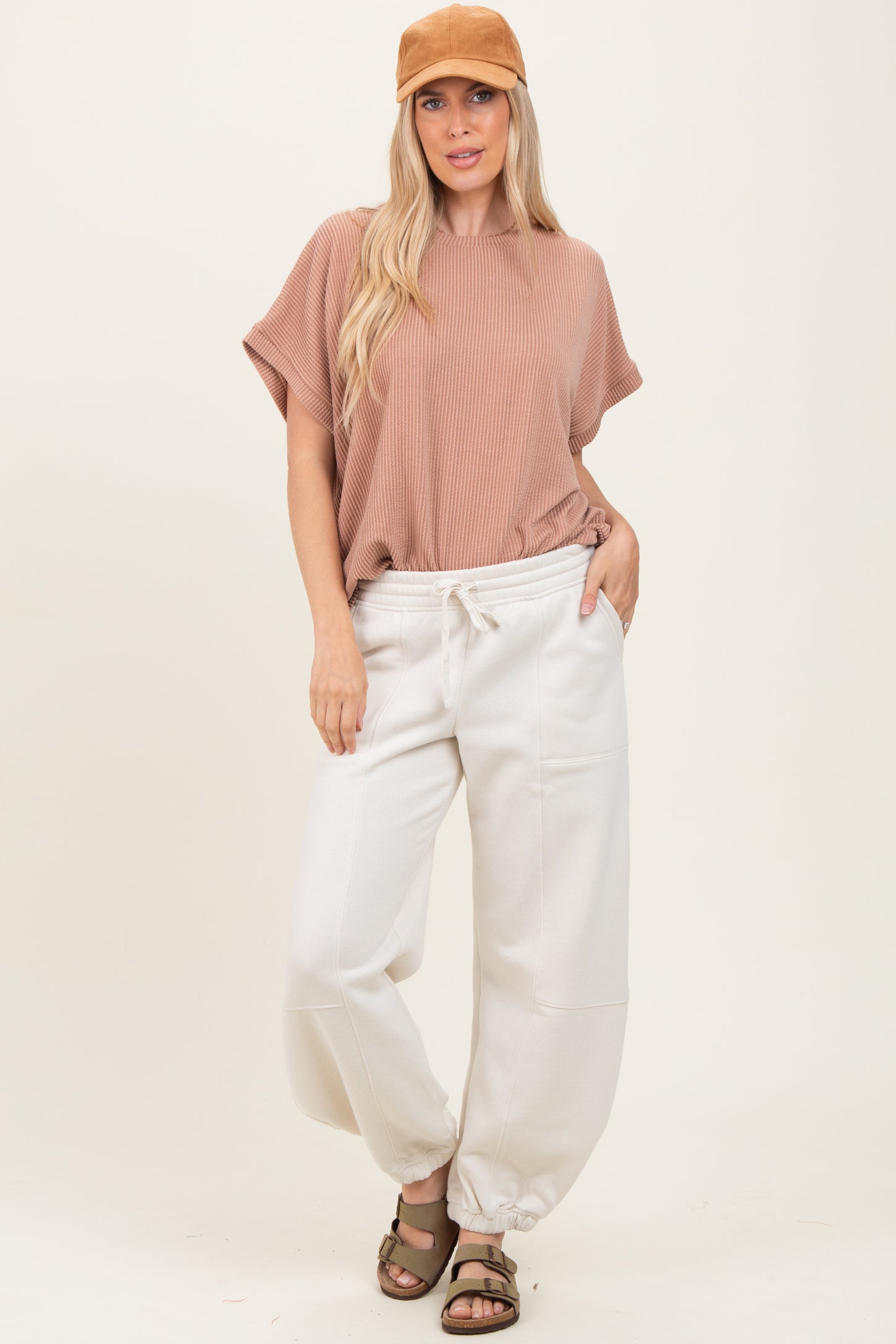 woman wearing a beige striped oversized top and cream jogger pants, styled with a brown cap and sandals