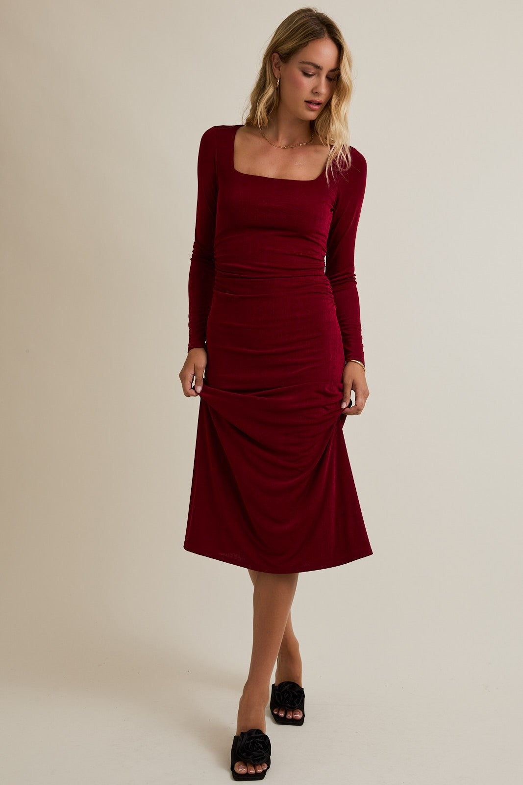 woman wearing a burgundy long sleeve ruched midi dress with a square neckline and styled with black slide sandals