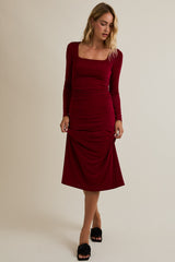 woman wearing a burgundy long sleeve ruched midi dress with a square neckline and styled with black slide sandals